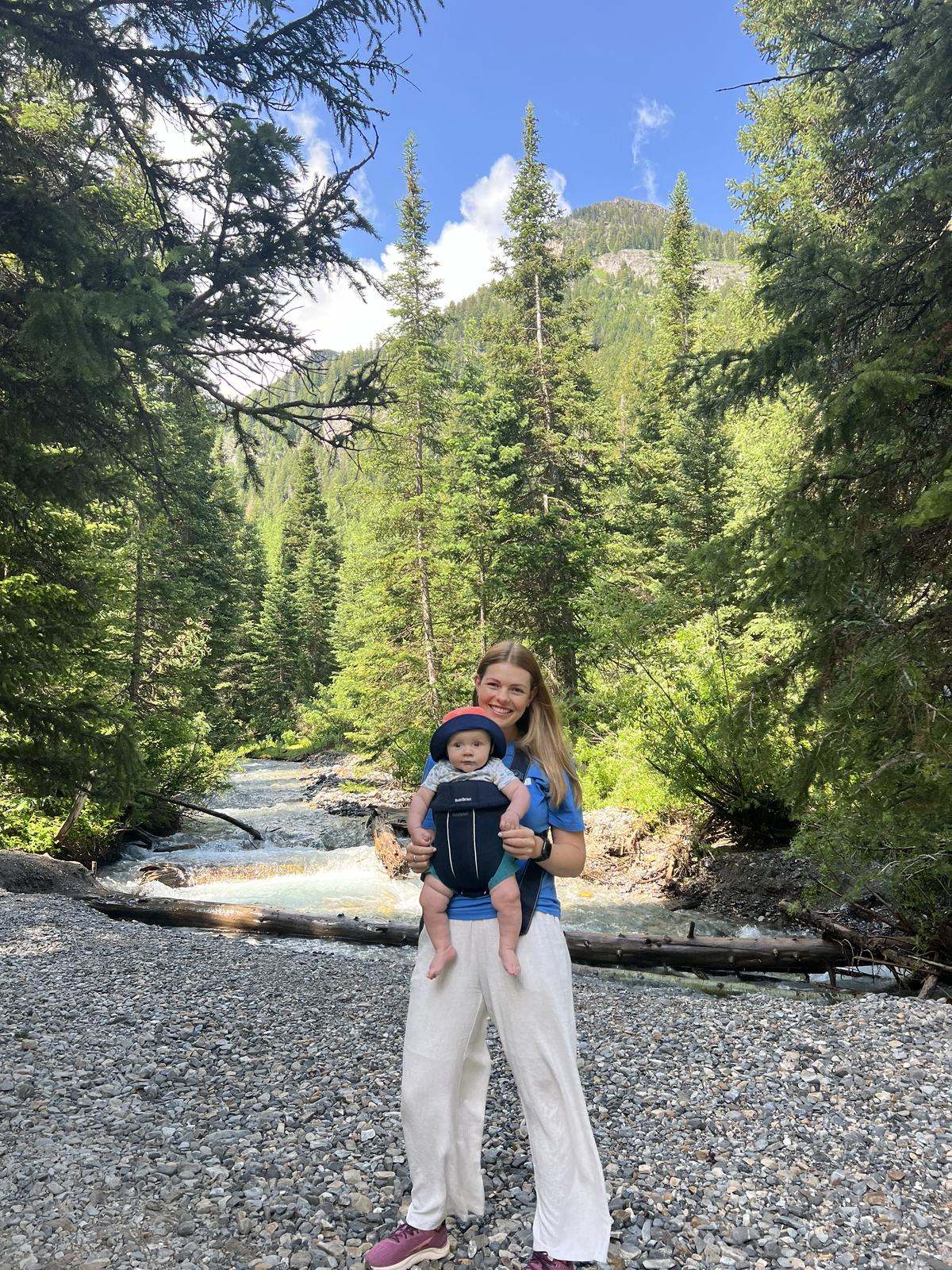 Allison in a blue shirt, holding a baby in a carrier beside a river in the pines.
